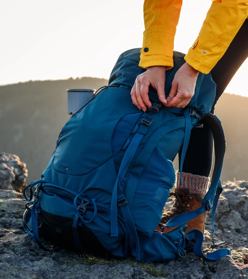 New Project - 2025-02-12T223646.945 Person in a yellow jacket adjusts the straps of a large blue hiking backpack on a rocky terrain, with a mountainous background.