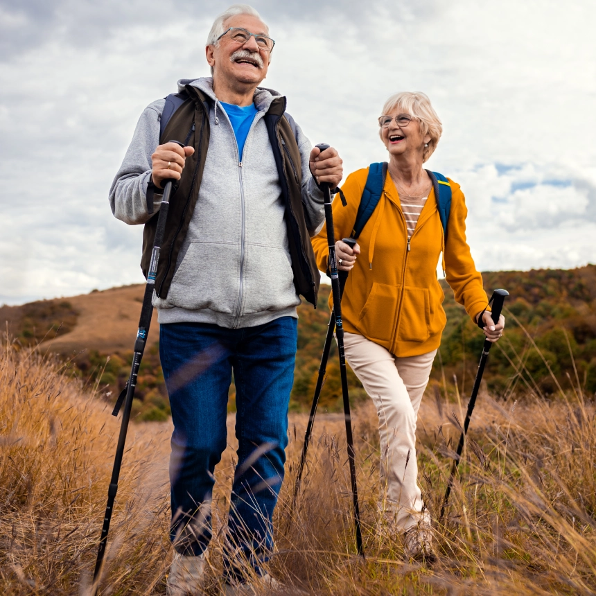 New Project - 2025-02-12T232237.978 Elderly couple hiking through grassy field with walking sticks, wearing backpacks and jackets, under a cloudy sky.