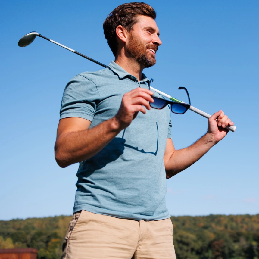 Man outdoors holding sunglasses and resting a golf club on his shoulder, wearing a blue polo shirt and beige shorts, with a blue sky and forested background.